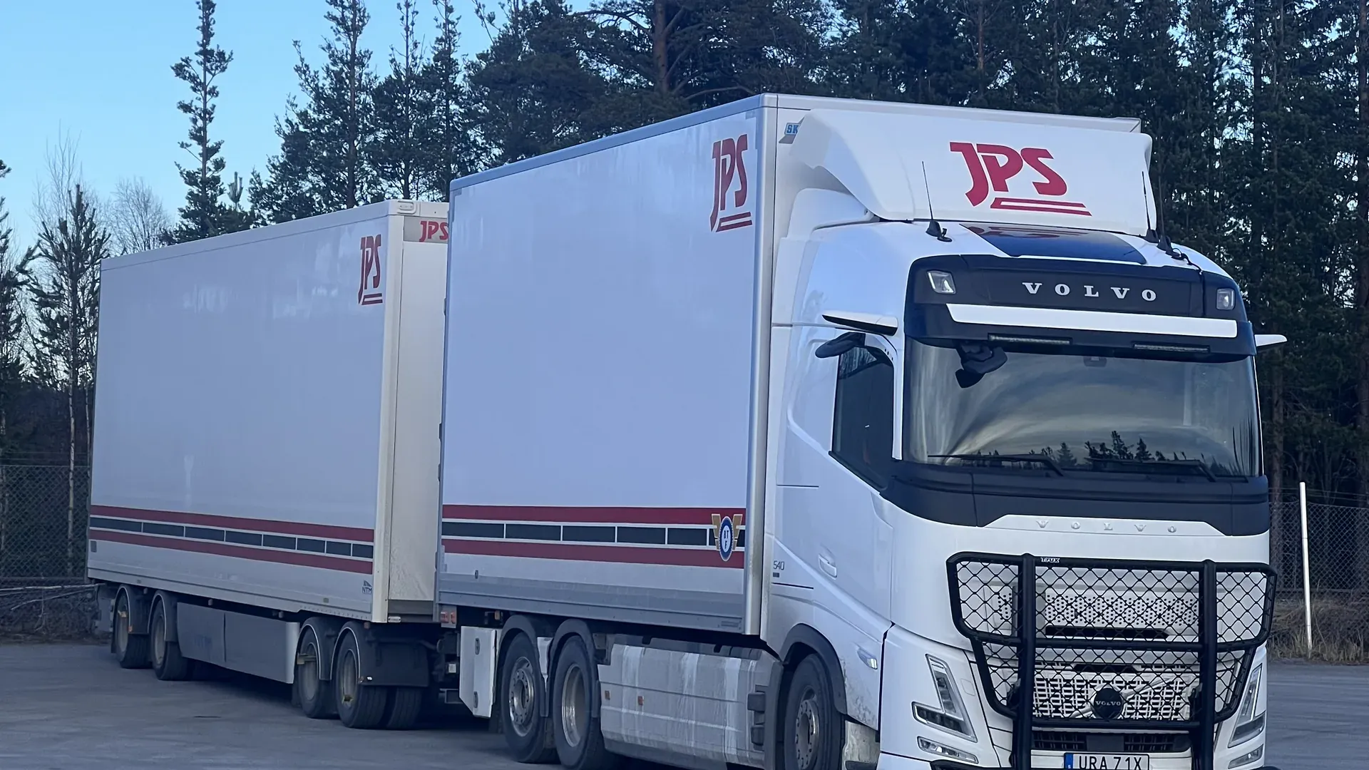Vit Volvo dragbil med två JPS-märkta släp uppställd på en grusplan med barrskog bakom.