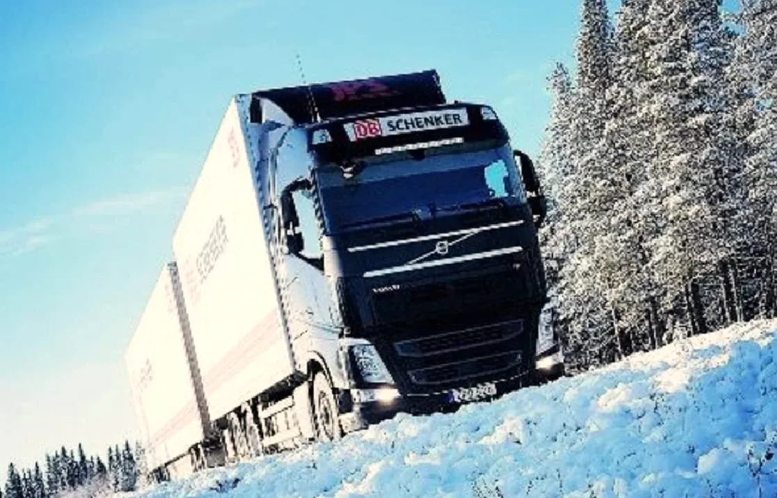 Volvo med skåp på vinterväg genom snötyngd granskog.